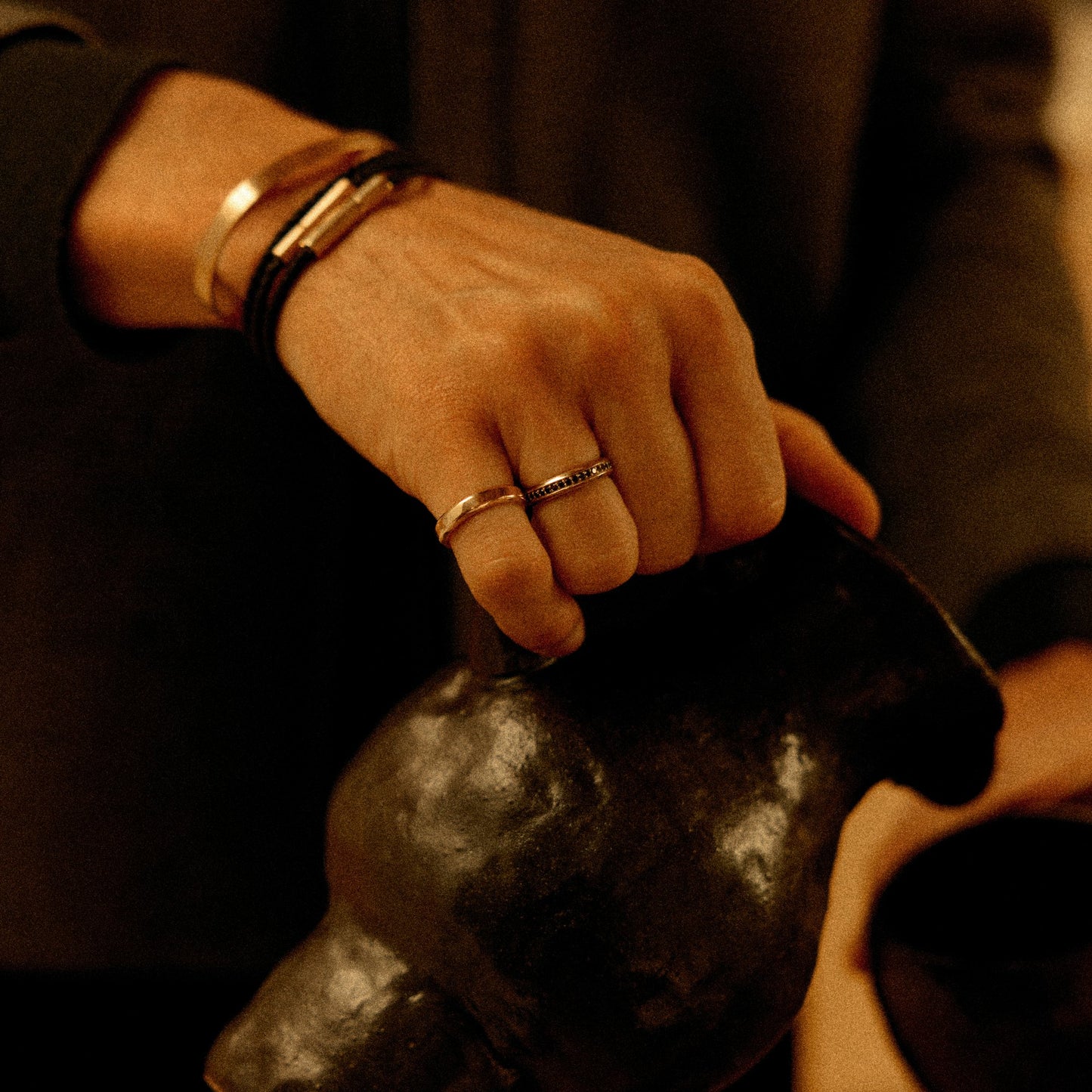 Hand holding ceramic jug wearing 18k gold cuff bracelet and black cord bracelet with 18k gold clasp