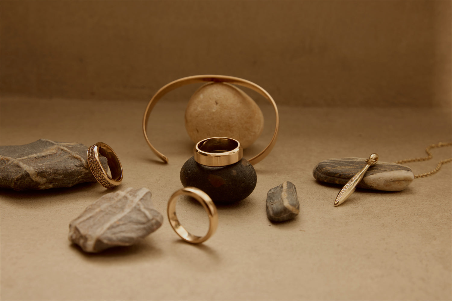Still life of 18k recycled gold cuff, rings and pendant arranged on stones.
