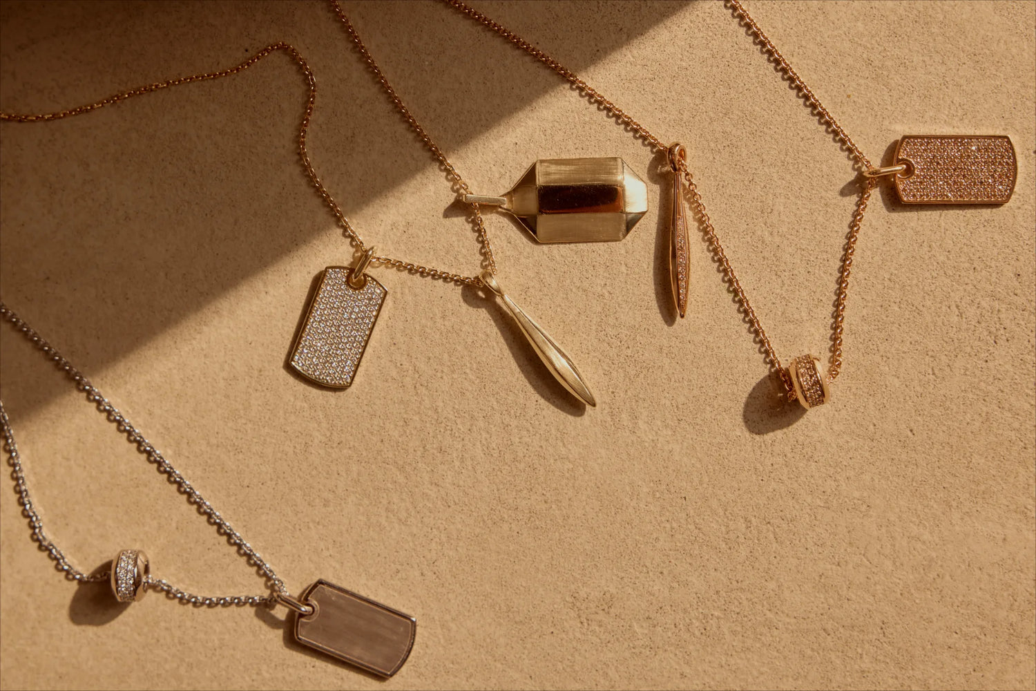 Flat-lay hero of mixed pendant necklaces in solid 18k recycled gold on a warm sand backdrop with natural shadows.
