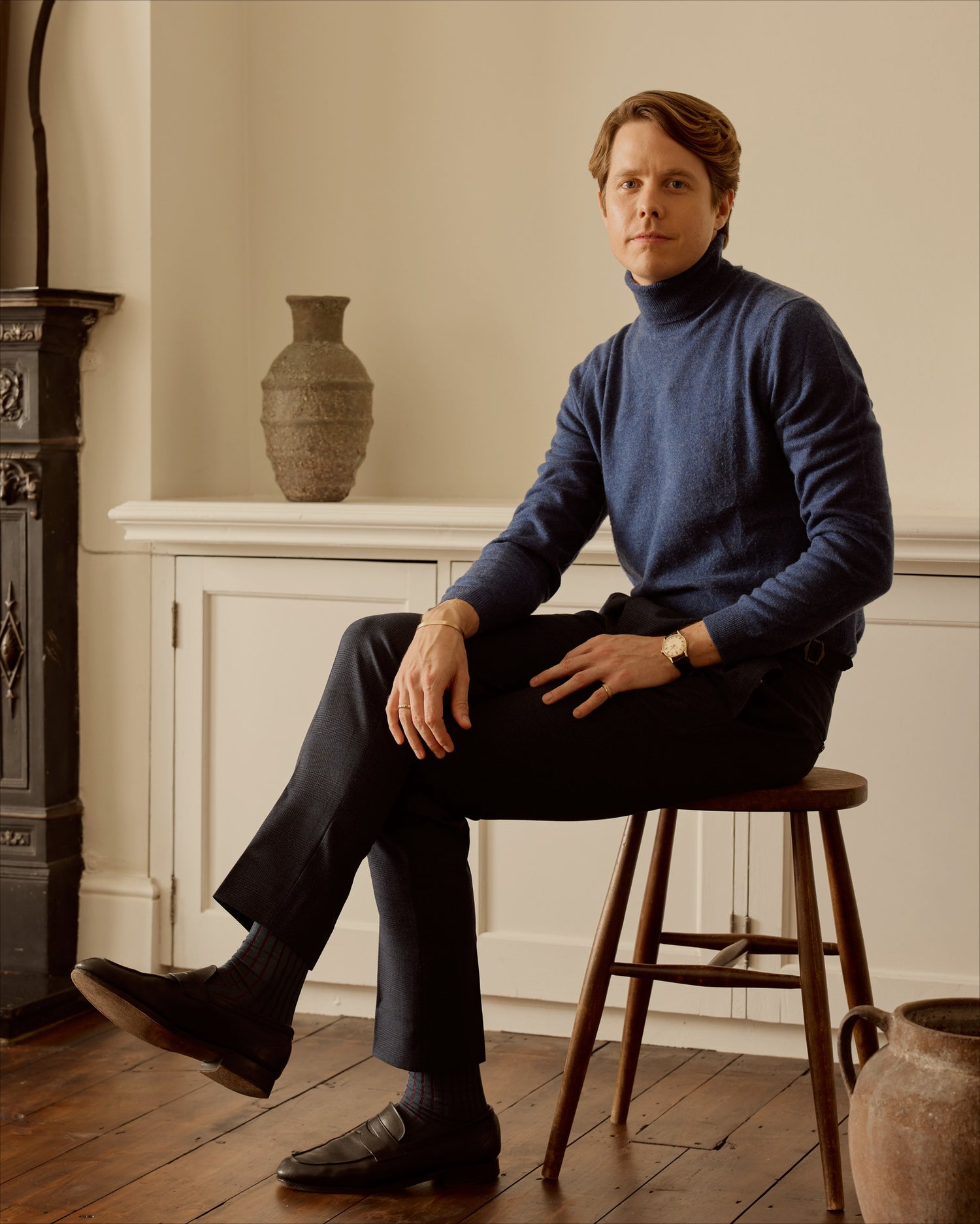Man seated on a stool in a blue turtleneck in a room with a fireplace and decorative vases, wearing an 18k gold cuff and band ring.
 
