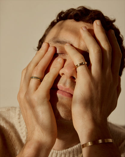 Portraitof man with hands covering face wearing pavé and classic 18k gold rings plus a slim cuff.
