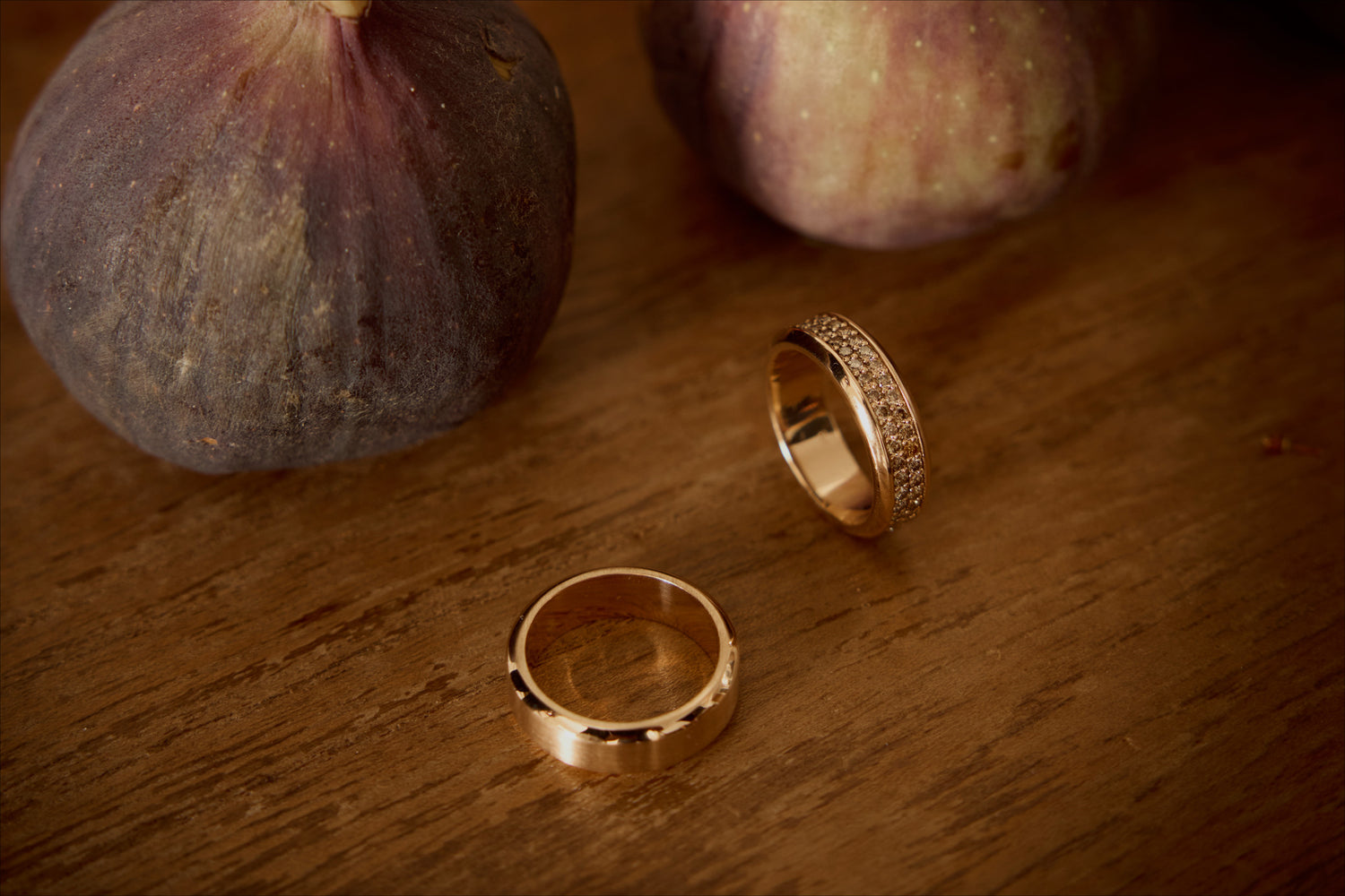 Two 18k recycled rose-gold bands on wood counter with figs in background. One pavé set, one classic polished.
