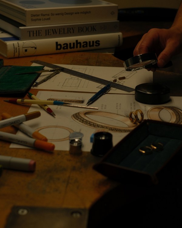 A hand holding a loupe over a dimensioned drawing, with a steel rule, compass, mechanical pencils and an instrument case around it, and gold rings resting on the sheet as references.