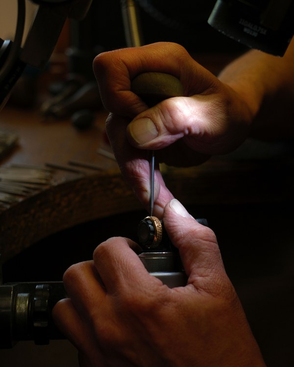 The heel of a palm driving a graver, its tip on a grain-set band held in a setting clamp, with a rack of gravers and burrs at the edge of the bench.