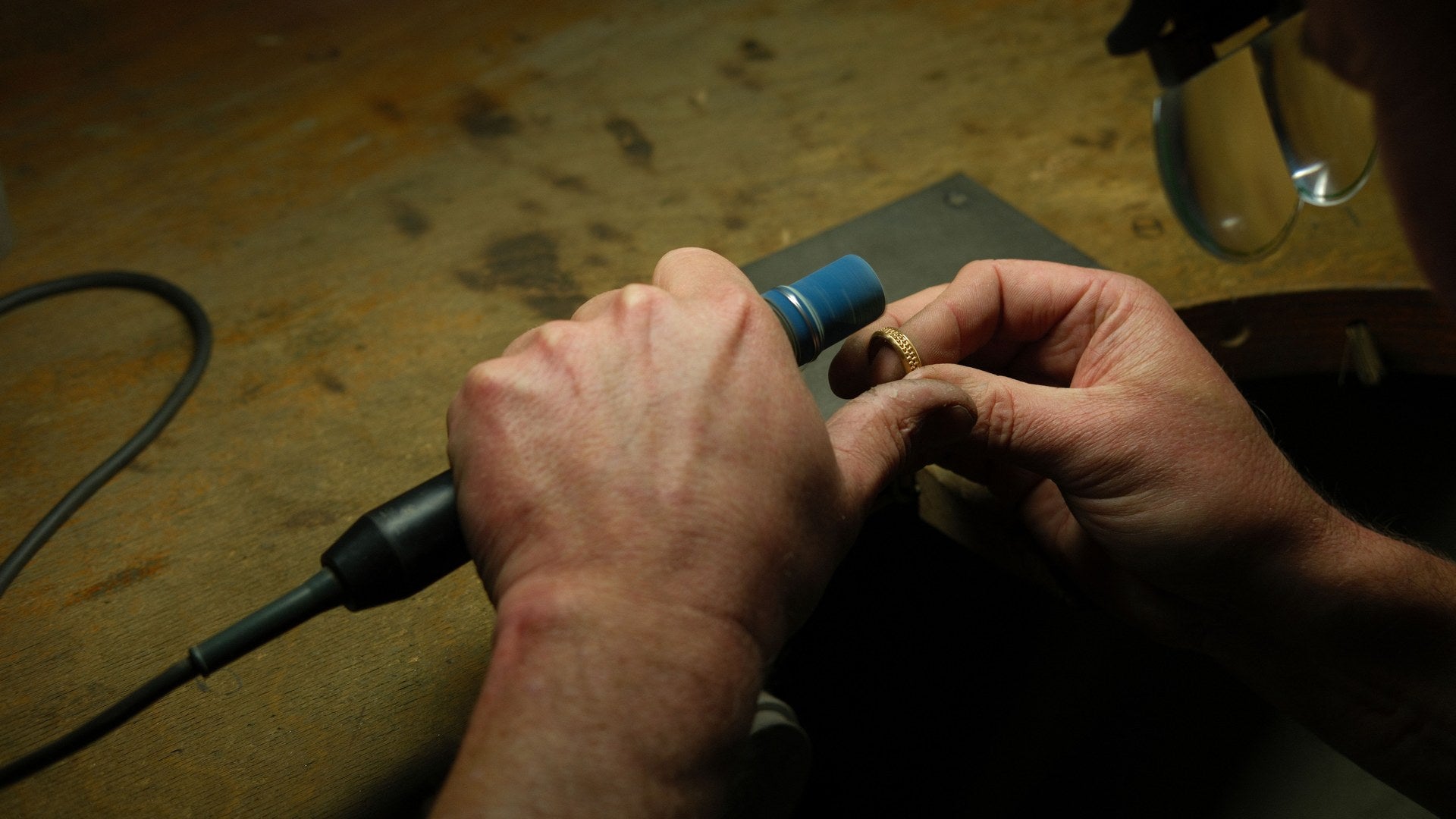 Two bare hands at a worn wooden bench, holding a gold band against a rubber holder on a pendant-motor handpiece, with the bench pin and swarf tray alongside.
